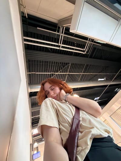A low-angle, rotated, medium shot shows a person with red hair wearing a beige shirt and black shorts, with a burgundy belt visible. The person is posing with their face close to the camera. The background features industrial-style architecture including metal beams, pipes, and ventilation structures. The location appears to be an interior space with a bright ceiling. The context suggests the scene might be in the city of Lautzenhausen, Germany.