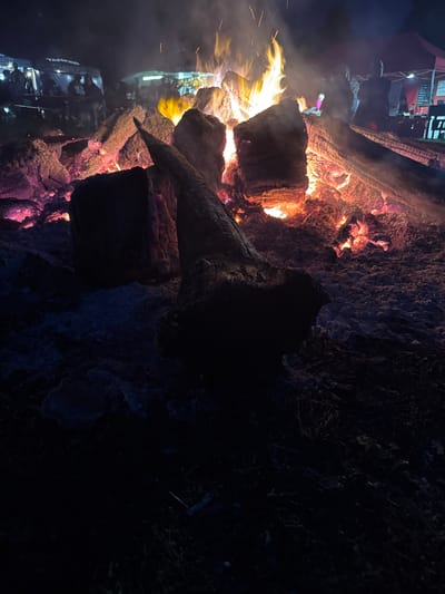 The image shows a large bonfire at night, likely at an outdoor event or gathering. In the foreground, several large logs are burning, with flames and embers illuminating the scene with orange and pink hues. Some of the logs are charred and broken, with a particularly prominent, dark, pointed piece of wood in the center. The ground around the bonfire appears to be covered in ash and possibly some snow or frost, creating a textured, dark surface.
In the background, silhouetted figures can be seen gathered under tents and canopies. These figures suggest the presence of a public event, possibly a festival or market, given the structures that resemble vendor stalls. The lighting from the bonfire casts a warm glow on the scene, contrasting with the dark night sky. The overall atmosphere is one of warmth and community, centered around the large fire. While the image itself does not explicitly state the location, the user prompt asks for a description of the location in Rapla, Estonia. Without additional context or visual cues within the image, it's impossible to definitively confirm this specific location.