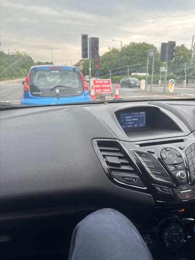 The image shows the interior of a car stopped at a traffic light in Lancaster, UK, at 54.07 latitude and -2.779 longitude. A blue Peugeot car is stopped directly in front of the car. The traffic light is red. There is a road sign in front of the Peugeot that reads: "When red light shows wait here". The driver’s seat is occupied by a person wearing blue jeans. The car's dashboard shows a screen displaying the BT audio system. The display shows a song called "Agony" by Sleepy Hollow, playing at track number 13 out of 14. The dashboard has a radio, CD player, AUX, phone, and menu buttons. The car has a black interior and a black steering wheel. The driver’s side window is dirty and has scratches. There is a red traffic light visible in the background.