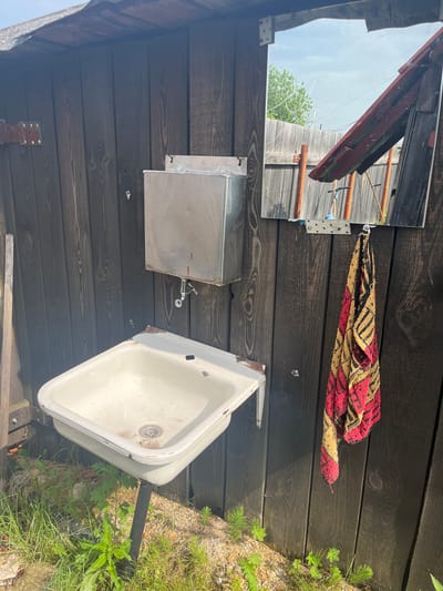 The image depicts an outdoor sink and a mirror mounted on a dark wooden wall. The sink is white, porcelain, and appears aged, with rust spots around the edges and faucet. A metal water tank is mounted above the sink, with a silver faucet attached. A towel with a red, yellow, and black striped pattern hangs from a hook to the right of the mirror. The mirror itself is rectangular and reflects an outdoor scene with a wooden fence, trees, and a cloudy sky. The area around the sink is overgrown with grass and weeds, suggesting it's in a rural or neglected outdoor setting. Based on the provided information, this scene is located in the city of Berezovskii, Russia.