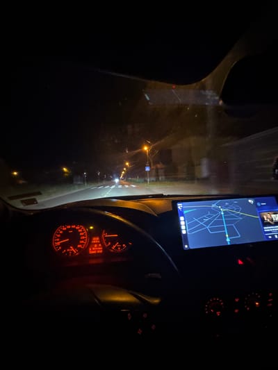 The image depicts the interior of a car at night, likely in transit. The dashboard is visible, with illuminated gauges showing a speed of approximately 15 km/h, and the time is 23:32. A navigation screen displays a map, possibly guiding the driver. The environment appears to be a road with streetlights. The OCR text suggests the car is in the vicinity of "KONSTANTIN & MAR," and a distance of "10.4 1200km" is displayed. No people are visible. Given the information, it's plausible the car is in or near Mogila, Bulgaria, based on the identified location on the navigation screen.