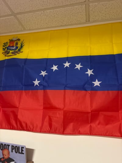 The image shows the flag of Venezuela hanging vertically. The flag features three horizontal bands of yellow, blue, and red. Eight white stars are positioned in an arc across the blue band. The yellow band displays the Venezuelan coat of arms on the bottom left. The flag is slightly wrinkled. A drop ceiling with white tiles can be seen on the left. Part of a book titled "FOOT POLE" is visible on the right, near a light-colored wall.
