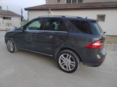 This media file shows a dark grey Mercedes-Benz SUV parked on a concrete surface in what appears to be a residential area. The car is viewed from its rear three-quarter angle, with the driver's side and rear end prominently featured. The vehicle has alloy wheels, tinted windows, and a roof rack. The background consists of buildings with tiled roofs and weathered walls, suggesting a rural or older urban setting. One building has a window with bars. A chain-link fence is visible behind one of the buildings. The sky is overcast, and the lighting suggests it is either early morning or late evening. There is no visible text in the image. The scene appears to be peaceful and static, with no activity or people present. The location context is Ignatievo, Bulgaria.