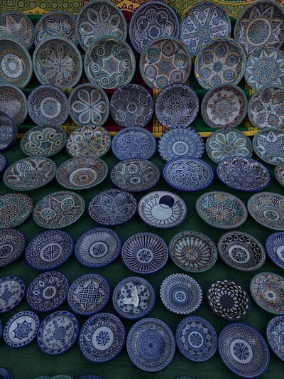 The image shows a collection of decorative plates, likely arranged for sale or display. The plates are predominantly ceramic and feature intricate patterns, with color schemes including blue, white, and earth tones. The arrangement appears to be on a green surface that may be artificial turf, although some plates rest on a decorative fabric at the upper left edge. The plates' designs vary but share a common theme of geometric and floral motifs, often radiating from the center. The presence of the plates suggests a marketplace or a retail environment that specializes in decorative art.

Given the information about the city of Burjassot, Spain, the plates may be local crafts for sale in that city. The city and specific location name are not represented in the image, however.