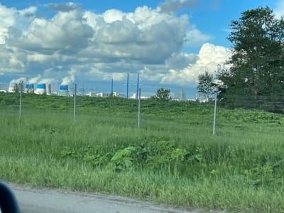 The image captures a view from the outskirts of Novodrozhzhino, Russia. A field of green grass occupies the foreground, with a wire fence running across it. In the background, several industrial cooling towers with blue and white markings are visible. High-voltage power lines and additional buildings suggest an industrial area or power plant in proximity to the town. A stand of trees is located on the right side of the image. The sky is partly cloudy with a gradient from light blue to white.