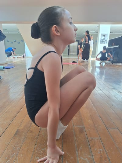 In a dance studio with wooden floors, a young girl in a black leotard is practicing a yoga pose. Her legs are drawn up to her chest, and her arms are wrapped around them. Her head is tilted back, and her eyes are closed, suggesting concentration or relaxation. In the background, reflected in a large mirror, several other dancers are also practicing. Some are standing, while others are in similar poses. A piano is visible to the left, and a few framed pictures hang on the wall. A man in a blue shirt and dark pants is seen helping a young boy with a pose. The lighting suggests it is daytime.