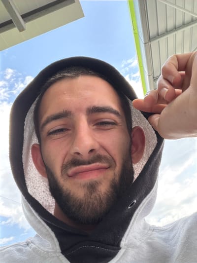 The image shows a man with a beard, looking down at the camera. He is wearing a white hoodie with a dark gray lining around the hood. The sky is visible in the background. The man's hand is visible adjusting the hood. The presence of both the sky and possibly roof-like features suggests the photo may be taken outdoors, potentially in Yoakim Gruevo, Bulgaria, though concrete confirmation of the location is absent from the image.
