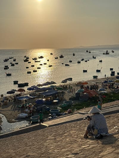 The image displays a coastal scene at sunrise or sunset in Thành phố Phan Thiết, Vietnam. The sun, a bright disk, is high in a hazy, golden-orange sky, casting a strong reflection across the calm ocean. Numerous fishing boats, including smaller circular basket boats, are scattered across the water, many appearing as silhouettes against the sun's glare.
On the sandy beach, a market operates with multiple temporary stalls and shelters, identifiable by various colored tarps and umbrellas (blue, green, orange, striped). Numerous individuals are gathered around these stalls, indicating commercial activity. Large circular basins are present on the sand near the market area.
In the foreground, a textured, sloped stone or brick embankment descends towards the beach. A person wearing a traditional conical Vietnamese hat (nón lá), a light-colored long-sleeved shirt, and patterned pants sits facing away from the viewer on this embankment. They are holding and observing a smartphone. A small woven bag is slung over their shoulder, and a clear plastic cup rests nearby. To the left, another person in a green shirt walks down the embankment. A third individual, possibly a child, stands near the seated person on the right.