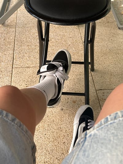 The image displays a first-person, downward perspective of an individual's lower body in an outdoor or semi-outdoor setting within Vandœuvres, Switzerland. The subject wears light blue denim shorts, white athletic socks, and black and white Vans sneakers with white laces. The left sneaker has its tongue folded over the laces, secured by a white fabric band bearing a "Vans" logo. The left foot is positioned with the shoe resting on a lower horizontal bar of a black metal stool or chair, which features a round black seat. The right foot is also visible, resting on the ground. The ground surface is composed of light-colored, rectangular paving stones or tiles. A light-colored furniture element is partially visible in the upper left corner. The scene is illuminated by natural light.