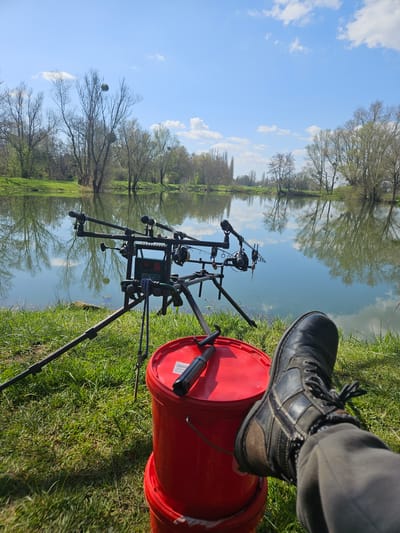 A fishing scene unfolds in Gavisse, France, beside a tranquil body of water reflecting the sky and surrounding trees. A fishing rod stand, equipped with multiple fishing rods, sits poised at the water's edge. Adjacent to the stand, a red bucket is positioned on the grass. A person's leg and boot rest atop the bucket.
