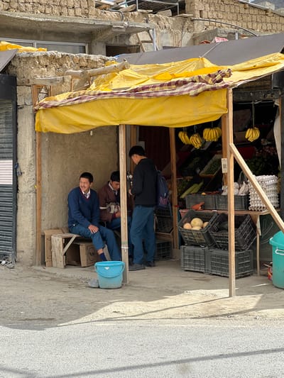 The image shows a street market stall in Yogma Mangmore, India. The stall is covered by a yellow tarp and sells a variety of fruits and vegetables. Three young men, appearing to be students in uniform, are present. One young man sits on a wooden bench, smiling and looking to the left. Another young man stands behind him, partially obscured. A third young man stands to the right, facing away from the camera, holding a mobile phone. In the foreground, a blue bucket and some rocks are on the ground. Crates of produce, including bananas, squash, and eggs, are visible within the stall. The background shows a stone building under construction.
