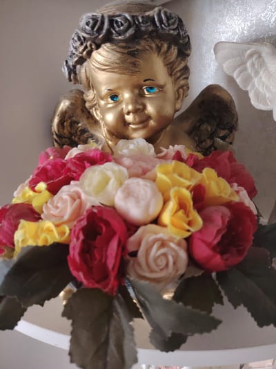 This is a medium shot, eye-level, close-up photograph of a gold-colored cherub figurine holding a bouquet of artificial roses. The cherub has blue eyes and a wreath of dark roses on its head. Its wings are also visible. The bouquet is composed of roses in various colors including red, pink, yellow, and cream. Dark green artificial leaves are interspersed with the roses and surround the base of the bouquet. The background is a plain, light-colored wall. To the right of the cherub, a portion of a white decorative element, possibly another figurine or architectural detail, is visible. The lighting suggests it is daytime. The overall impression is that of a decorative item, possibly for a home or a special occasion. No text is visible in the image. The location context provided is Razvilka, Russia.