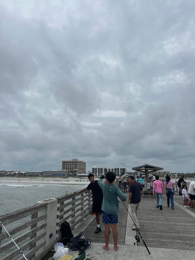 Here's a concise description of the image:
The image shows a section of a pier with people fishing. The pier's wooden structure is visible, and people are standing along the edge, some with fishing rods. In the background, the beach and the ocean are seen, with buildings along the coastline. The sky is cloudy.
The image appears to have been taken at the Jacksonville Beach Pier.