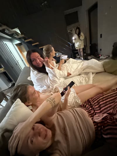 Four people, three women and one person of indeterminate gender, are lying on a bed or couch, seemingly in a relaxed setting. The image appears to be taken indoors, possibly in a bedroom or living room. One woman on the far right is holding a remote control and looking at the camera with a smile, her hand covering part of her face. Next to her, another woman is lying down, with a tattoo visible on her arm. In the center, two more people are lying down, one wearing a white robe and the other with blonde hair. The lighting suggests it might be evening or a dimly lit room. The scene conveys a sense of casual comfort and togetherness. There is no visible text in the image.