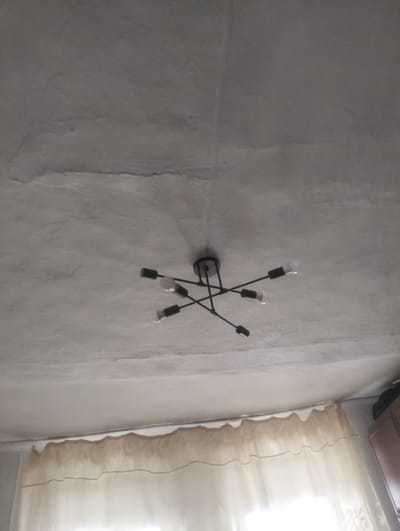 The image shows a ceiling with a modern black chandelier. The chandelier has multiple arms extending from a central point, each with a light bulb. The ceiling appears to be textured and possibly aged, with some visible lines or cracks. Below the ceiling, a translucent curtain is visible, suggesting this is an indoor scene. The lighting suggests it is daytime. There are no people or animals present in the image. The location in the city of Qarabulaq, Kazakhstan is not evident from the visual information provided in the image.