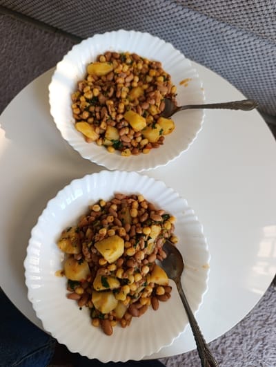 Two white, fluted ceramic bowls are positioned on a round, white table surface. Each bowl contains a serving of Githeri, a Kenyan dish comprising light brown beans, yellow corn kernels, diced potatoes, and chopped green herbs in a reddish-orange sauce. A silver-colored spoon with an ornate handle rests within each bowl. A partial view of blue denim fabric, consistent with a person's jeans, is visible in the lower-left foreground. The background features a textured, light grey fabric surface. The scene is located within the Kahawa Wendani ward, Kenya.
