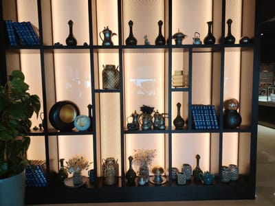 The image shows a dimly lit interior display shelf unit, likely in Konigil, Uzbekistan. The shelves are dark wood with illuminated back panels. The unit contains various decorative objects, including books, vases, teapots, bowls, and what appear to be decorative lanterns. A potted plant is visible on the far left. The objects are arranged symmetrically on each shelf. The overall impression is one of an upscale retail or hospitality setting, showcasing artisanal or antique items. There are no people visible in the image.