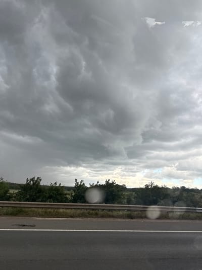 The image captures a scene from a moving vehicle, likely on a highway, looking out towards a rural landscape. The dominant feature is the dramatic, heavily clouded sky, characterized by dark, swirling grey clouds that suggest impending rain or a storm. Below the sky, a line of dense green trees and foliage forms the horizon, with patches of lighter green fields visible behind them. A metal crash barrier separates the road from the verge, which is covered in dry grass and some scrub. The foreground consists of a dark asphalt road with a white line marking the lane. Several circular, blurry white spots are present on the glass through which the photograph was taken, possibly water droplets or reflections. The overall impression is one of a moody, overcast day in a semi-rural setting. The location is specified as Hertsmere, United Kingdom.