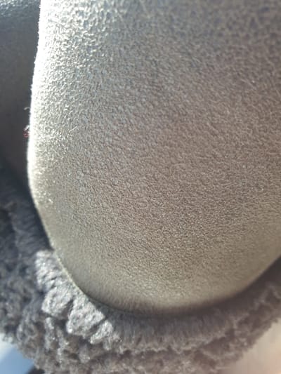 The image displays a close-up, out-of-focus view of two distinct textile materials. The prominent upper and central portion features a light brown to tan-colored material with a finely napped texture, consistent with suede or nubuck. This material exhibits a convex, curved form. Strong directional lighting from the upper-left creates highlights that accentuate the granular surface texture. Adjoining the lower and left edges of this material is a dark grey to black textile, characterized by a dense, tightly curled or looped pile, indicative of synthetic fleece or sherpa lining. No human figures, specific actions, or extraneous environmental details are discernible within the frame. The location is Kinanie ward, Kenya.