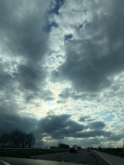 The image captures a view from a moving vehicle, likely on a highway, with the primary focus being the sky. The sky is filled with a mix of dark, heavy clouds and patches of brighter, sunlit areas. The light is breaking through the clouds in places, creating a dramatic effect. The highway is visible on the right side of the frame, with a guardrail, some trees, and several cars in motion. The scene does not indicate that the location is in the city of Gabarnitsa, Bulgaria.