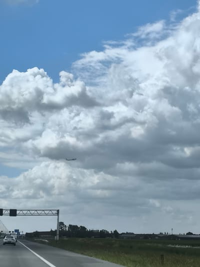 The image captures a highway scene near Hoofddorp, Netherlands, under a sky dominated by cumulus clouds, partially obscuring the blue sky. A white passenger car travels on the highway, with an overhead gantry spanning the road. In the distance, there are trees and low-lying buildings, likely industrial or agricultural structures, surrounded by fields. An airplane is visible in the sky, appearing small against the backdrop of the clouds. The road is marked with white lines, and greenery flanks the highway's edge.