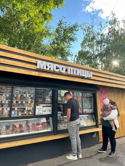 This image captures a street-level view of a small food shop with prominent Cyrillic signage that reads "МЯСО ПТИЦЫ" (Meat Poultry). The shop appears to be a butcher or deli, as the display windows are filled with various food items, including what looks like bread, sausages, and packaged meats, with price tags visible.
Two people are in the foreground. On the right, a person wearing a black t-shirt and grey pants is bending over, possibly looking at or interacting with the shop's display. On the left, a woman is carrying a child on her back. The child is wearing a light-colored jacket and a pink hat.
The shop itself has a modern facade with vertical wooden slats. Above the shop's entrance, there is a red sign with decorative white patterns. The overall scene suggests a sunny day, with dappled sunlight filtering through green trees that frame the top left of the image. The sky is blue with some white clouds. The ground in front of the shop appears to be asphalt or a similar paved surface.
Visible text on the shop windows includes price indications like "365" and "329-", and a sign that says "ПРОВЕРЯЙТЕ ТОВАР И ДЕНЬГИ, НЕ отходя ОТ КАССЫ" (Check goods and money without leaving the checkout). Another sign reads "АКЦИЯ" (Sale/Promotion) and "ОХЛАЖДЕННАЯ ПРОДУКЦИЯ" (Chilled Products).
The overall impression is that of a casual, everyday scene in an urban or suburban setting, likely during daytime.