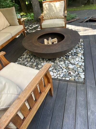 The image captures a tranquil outdoor seating area featuring a round, weathered metal fire pit surrounded by smooth, light-colored river stones. Two wooden lounge chairs with thick, cream-colored cushions are positioned around the fire pit, one on the left and another partially visible on the right. The setting is a dark wooden deck, suggesting a contemporary outdoor living space.

Behind the fire pit and to the left of the chairs, lush green foliage and trees are visible, hinting at a natural, possibly wooded or garden environment. Sunlight filters through the trees, casting dappled shadows on the deck, indicating it is daytime, likely late morning or afternoon. The overall mood is one of relaxation and comfort, inviting outdoor leisure. There is no visible text in the image. The setting appears to be a private backyard or patio.