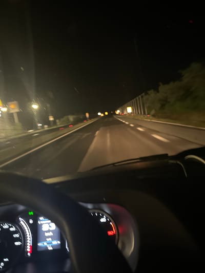 The image presents a sideways view from inside a car driving at night. The scene is dominated by darkness, typical of nocturnal conditions.
On the right side of the frame, a segment of the car's interior is visible, including the steering wheel and a clear view of the instrument cluster. The digital display shows "66 km/h," indicating the vehicle's speed. Below this, "14:48" is displayed, possibly representing the time of day or a trip duration, and "900.0 km" is visible, likely a trip odometer. A green indicator light is on, and a prominent red warning light is active near the '200' mark on an analog gauge, possibly indicating an issue with the engine or oil pressure. Another gauge shows a high red bar, which could be a fuel or temperature indicator.
The left and central parts of the image show the exterior, featuring a paved road with clear white lane markings. The road stretches into the distance, illuminated by the car's headlights and various distant light sources. Numerous bright yellow and red lights are scattered along the side of the road, suggesting streetlights, road reflectors, or the taillights of other vehicles. Dark forms of trees or foliage are barely discernible against the dark sky along the road's edge. A faint red light appears high in the top-left portion of the sky, possibly a distant aircraft or a light on a tall structure.
The image captures the activity of driving a car at night. The vehicle's speed is displayed, and the visible metric units align with the context of Hadzhidimitrovo, Bulgaria. The weather appears clear, with no rain or fog evident.