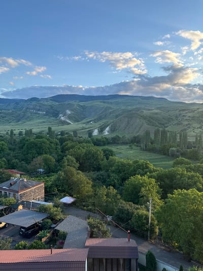 The image shows a landscape scene from the city of Aspindza, Georgia, likely taken from an elevated position. In the foreground, there are several buildings with tiled roofs and a stone-clad structure. A dark-colored SUV is parked under a carport. A road or pathway is visible, lined with trees and vegetation.
The midground is dominated by a dense forest of lush green trees. Beyond the forest, rolling hills stretch into the distance, displaying a patchwork of green fields and lighter-colored soil or rock formations. The hills are sculpted with numerous contours and valleys, indicating a natural, possibly eroded landscape.
The background features a range of mountains under a blue sky with scattered white and gray clouds. Sunlight is breaking through the clouds, casting rays across the sky and illuminating parts of the landscape. The overall atmosphere suggests a late afternoon or early evening setting, with soft, diffused lighting. The scene is tranquil and natural, with a mix of human habitation and wild terrain.
