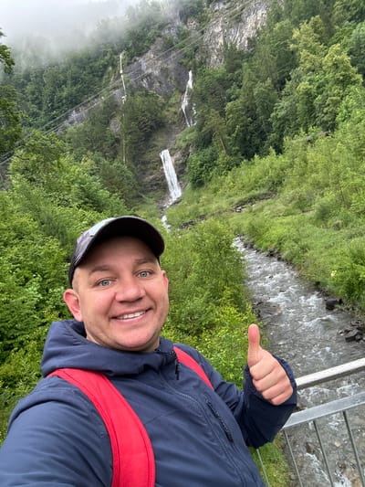 In a selfie taken outdoors, a smiling man with blue eyes and a baseball cap stands in front of a lush green natural landscape. He is wearing a dark blue hooded jacket with a red backpack over it, and he gives a thumbs-up gesture with his right hand. Behind him, a narrow, fast-flowing river runs through a rocky bed, flanked by dense green foliage. Further in the background, a tall, cascading waterfall plunges down a steep, rocky cliff face partially obscured by trees and mist. The overall scene suggests a scenic location, possibly in the Swiss Alps, given the mention of Hüsen, Switzerland. The environment is characterized by natural elements like water, rocks, trees, and a waterfall, with a slightly overcast or misty sky contributing to the atmospheric setting.
