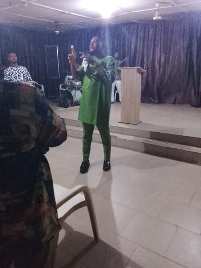 A man in a green suit stands on a stage with a podium, gesturing with his hands. Other people, including musicians with instruments, are visible in the background. The lighting is dim, and the scene appears to be in a room or hall with tile flooring. A white chair and part of a person’s body obstruct the view in the foreground. The backdrop is a dark curtain.