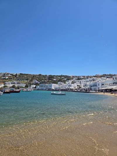 This is a landscape photograph taken from a slightly elevated perspective, showing a picturesque coastal scene. The image is oriented vertically, with the sky occupying the left portion and the sea and shoreline on the right.
The sky is a vibrant, clear blue, with a few distant birds visible. Below the sky, a densely built hillside town, characteristic of Mykonos, Greece, cascades down towards the sea. The town is composed of numerous white-washed buildings with red-tiled roofs, nestled closely together and dotted with greenery.
The sea is a brilliant turquoise, appearing calm and shallow near the shore, where the sandy bottom is clearly visible through the clear water. The gentle waves lap onto a golden sand beach. Along the waterfront, several boats are docked, including a few small wooden vessels and a larger white boat.
The shoreline is lined with buildings and people. Numerous individuals are seen walking along the beach and sitting at outdoor cafes or restaurants that appear to be built along the waterfront. The sunlight suggests it is a bright, sunny day, likely midday or early afternoon. The overall impression is one of a lively, idyllic vacation destination. No visible text is present in the image.