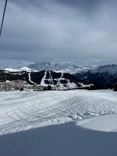 An elevated view captures a snow-covered mountain landscape in Villard-sur-Doron, France, under a partly cloudy sky. The foreground features an expanse of white snow, heavily crisscrossed with numerous ski and snowboard tracks. A dark shadow extends across the lower right portion of this snowy surface. In the midground, a cluster of buildings, consistent with a small town or village, is visible in a valley to the left. Beyond the village, dark green coniferous forests cover hillsides, interrupted by several distinct white ski slopes running down them. The far left edge of the image shows a dark, vertical cable, consistent with a ski lift mechanism. The background is dominated by a jagged range of snow-capped mountains stretching across the horizon, partially obscured by low-hanging clouds. No individuals or active interactions are visible.