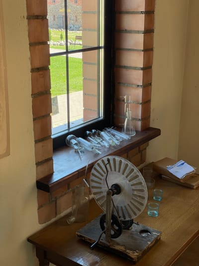 The image depicts a windowsill displaying a collection of laboratory glassware, including test tubes and a flask, set against a brick wall. Through the window, a grassy area with some stones and a bench is visible, with a stone building in the background. On the wooden table in front of the window, a spinning disc apparatus, a glass bottle with tubes, and two small glasses are arranged. A stack of books wrapped in brown paper with a white cloth on top rests on the table to the right. The scene suggests an indoor setting possibly within a historical or educational establishment in Літвенскі сельскі Савет, Belarus, given the architectural style and the presence of scientific apparatus. No people are present in the image.