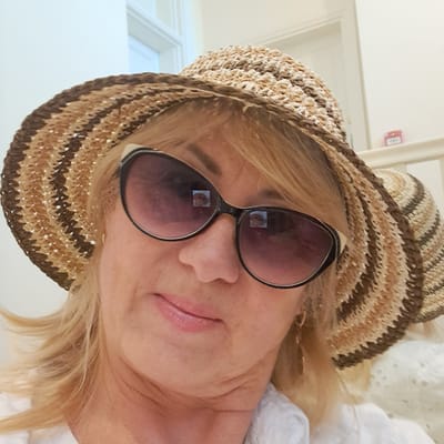 This media file shows a close-up, rotated portrait of a blonde woman wearing a straw hat and sunglasses. She is looking towards the camera with a slight smile. The sunglasses have a dark, reflective lens, and the frames are black and white. Her hair is blonde and appears to be layered. She is wearing a white top, possibly made of a light, breathable fabric with eyelet detailing. The background is out of focus, but appears to be an indoor setting with white walls and what looks like a doorway or window frame on the left. There is a small red fire alarm on the wall in the upper left corner. The lighting suggests it is daytime. The overall mood is relaxed and casual. The media file does not contain any visible text, apart from a small, blurry reflection of a person in the sunglasses.