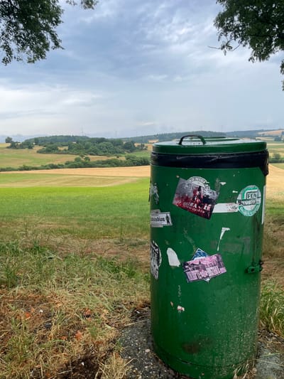 A green metal trash can is positioned on the right side of the frame, slightly angled towards the viewer. It is adorned with several stickers. In the background, a rolling landscape unfolds under a cloudy sky. The landscape features fields of green and golden crops, interspersed with patches of trees and rolling hills. Several wind turbines are visible in the distance, suggesting an open, rural setting. The foreground shows dry grass and a portion of a paved surface. The image appears to be taken outdoors in a natural environment, possibly on the outskirts of Maar, Germany.