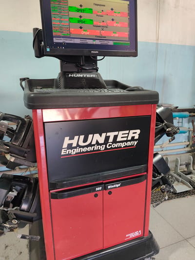 The image shows a Hunter 560 WinAlign wheel alignment machine in a repair shop. The machine is red and black, with a monitor displaying alignment data. The screen shows readings for camber, caster, and toe angles for a vehicle. The brand name "HUNTER Engineering Company" is visible on the side of the machine. The environment appears to be a workshop with tools and equipment in the background, including a car lift. The overall scene suggests a professional automotive service setting. No specific location in Atameken, Kazakhstan can be determined from the image.