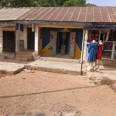 The image shows the exterior of a building located in Kufang, Nigeria. The building has a corrugated metal roof and is painted blue and yellow. There are two mannequins in front of the building, one wearing a blue dress and the other wearing a red and black dress. There is a small concrete sidewalk in front of the building, and the ground is covered in gravel. The scene is in a residential area of Kufang, Nigeria, with a few trees in the background. The image was taken during the day. The sun is shining and the sky is clear. The photo is taken at a wide angle, showing the front of the building from a distance. The photo is taken at a low angle.