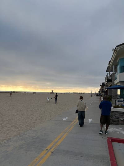 Here's a detailed description of the image:
**Location:** This image appears to be taken in Newport Beach, California, United States of America. The presence of the beach, buildings along the boardwalk, and the architectural style suggest this location.
**Scene:** The photograph depicts a beach scene during the day with cloudy skies. There's a stretch of sandy beach alongside a concrete boardwalk. Buildings are visible on the left side of the boardwalk.
**People:**
* Two people are walking on the boardwalk. One is wearing a blue shirt and dark shorts, and the other has a tan jacket and jeans.
* There are several other people on the beach, some with dogs.
**Objects:**
* The boardwalk has yellow lines dividing it.
* There are white arrow markings on the boardwalk.
* A lifeguard tower is seen in the distance on the beach.
**Actions/Interactions:**
* People are walking along the boardwalk and the beach.
* Some people are walking dogs.
**Environment:**
* The sky is overcast.
* The beach is sandy.
* Buildings are visible along the boardwalk.
