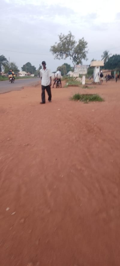The image depicts a street scene in what appears to be the city of Ichol, Nigeria. The ground is covered in reddish-brown soil, suggesting a dry or dusty environment. In the mid-ground, a man is walking towards the viewer, wearing a light-colored short-sleeved shirt and dark trousers. He is also wearing a dark baseball cap. To the left of the man, a person is riding a motorcycle with a passenger behind them, also traveling towards the viewer. In the background, there are several buildings, trees, and what seems to be a utility pole with a sign. Another motorcycle is visible in the right background, with a person on it. The sky is overcast with a light grey color, indicating diffused light conditions. The overall scene suggests a typical day in a Nigerian city, with daily activities like commuting by motorcycle and people walking on the street.