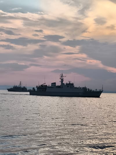 The image depicts two naval vessels on the sea under a cloudy sunset sky. The sky displays a gradient of colors, from light blue at the top to pink and purple hues lower down, with scattered grey and white clouds. The water is calm with gentle ripples reflecting the colors of the sky.
The foreground vessel, identified by the numeral "M271" on its hull, is a large grey naval ship. It is oriented diagonally from the lower right to the upper left of the frame, with its stern facing the viewer. Several figures, appearing to be crew members, are visible on the deck of this ship. The vessel has a prominent superstructure with various antenna and communication equipment.
Further in the background, to the left, is a smaller grey naval vessel, also moving on the water. This ship has a more slender profile and a raised mast. Distant land or mountains can be seen as a faint silhouette on the horizon.
The scene appears to be taking place at sea, likely during dusk or dawn, given the lighting and sky conditions. There is no indication of the city of Emona, Bulgaria, in this image, as the scene is solely focused on the naval vessels and the seascape.