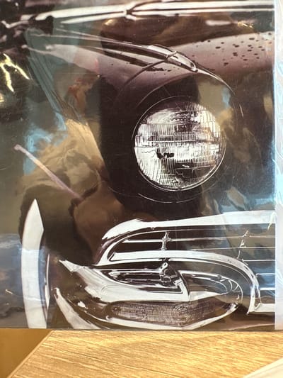 The image displays a close-up, angled view of the front right quarter of a dark-colored classic American automobile. The scene is located in Новоглаголево, Russia. A prominent circular headlight with a detailed fluted lens is centered in the upper-mid section. Below the headlight, a complex chrome grille structure with horizontal bars and vertical elements is visible, leading down to a wraparound chrome bumper. An amber-colored turn signal or parking light with a textured lens is integrated into the lower part of the bumper. The car's body features a dark, possibly black or deep blue, glossy finish, upon which numerous small water droplets are evident, suggesting rain or recent washing. Reflections of an unseen environment, featuring muted blue and brown tones, are visible on the car's painted surfaces and polished chrome. A chrome hood ornament or trim piece is present on the fender above the headlight. The vehicle's white sidewall tire is partially visible on the far left. The photograph itself appears to be a glossy print or poster, exhibiting surface scratches, dust, and significant reflections from an external light source, indicating it is covered by a protective layer or under bright lighting conditions. The bottom of the print rests on a light-colored wooden surface.