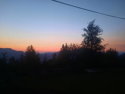 A twilight scene unfolds over the village of Slavsko, Ukraine, where the sky transitions from a pale blue at the zenith to a soft orange and pink near the horizon. Silhouetted against this colorful sky are rolling hills and a dense forest. Several trees, their branches reaching upwards, are prominent in the foreground, their dark forms contrasting with the fading light. A thin wire stretches diagonally across the upper portion of the image. The ground in the foreground appears to be a grassy area, with some indistinct objects scattered on it. The overall impression is one of quietude and natural beauty as day gives way to night.