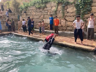 The scene depicts an outdoor water body in Killi Malik Sher Mohammad, Pakistan. In the foreground, two individuals are actively entering a clear, light blue-green pool. One individual, wearing dark clothing, is captured mid-air diving head-first, creating a significant splash. Approximately 18-20 males, ranging from boys to young adults, are present in various states of dress. Several young men, some in wet clothing like white tank tops and dark pants, stand barefoot on the wet, reddish-brown paved edge surrounding the water, observing the activity. One young man in a white tank top and dark pants is visibly adjusting his pants on the edge. The background features a rough-hewn stone wall, partially covered with green vines and foliage. Additional individuals are visible on the upper sections of this wall and an elevated dirt path beyond it, observing the scene below. The environment is brightly illuminated by natural sunlight.
