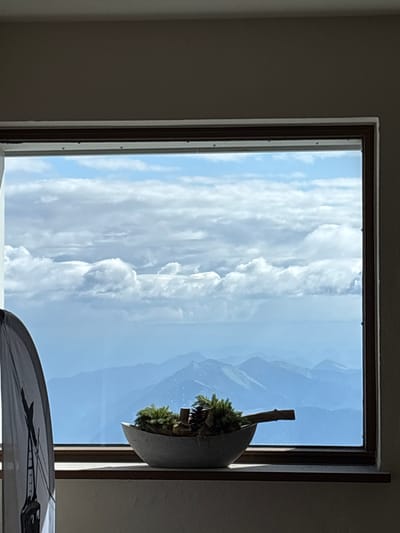 The image shows a view from a window in Ehrwald, Austria, overlooking a layered mountain landscape under a cloudy sky. In the foreground, on the windowsill, is a decorative bowl filled with greenery, pinecones, and a wooden stick. To the left of the bowl, partially visible, is a tall, white object with a black graphic, possibly a flag or banner. The window frame is brown, and the wall around it is a neutral, light brown color. The scene captures a serene and expansive view of the Austrian Alps.