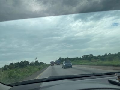 The image shows a view from inside a vehicle on a highway in Kwali, Nigeria. The sky is overcast with gray clouds. The highway is paved and has a yellow line marking the lanes. Several vehicles are on the road, including a blue hatchback and two trucks, one of which has a covered cargo. Lush green vegetation and trees line the sides of the highway, with some signs of human habitation in the distance, such as utility poles. The overall scene suggests a typical day of travel on a highway in a tropical region.