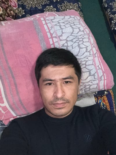 A frontal close-up depicts an adult male positioned indoors in Baygundi, Uzbekistan. The subject has short dark hair, a mustache, and light stubble. He is wearing a dark, long-sleeved crew-neck shirt, which bears a subtle, stylized Z-shaped logo on the left chest. The man is in a reclining posture, with his head resting on a pink pillow. The pillow displays grey vertical stripes and a white abstract pattern resembling cracked ground. Adjacent to the pillow on the right, a textile with traditional blue, yellow, and red geometric patterns is partially visible. Behind and above the pillow, a dark blue fabric with lighter floral designs is present, alongside a dark green floor covering to the right. The man maintains direct eye contact with the camera, exhibiting a neutral facial expression.