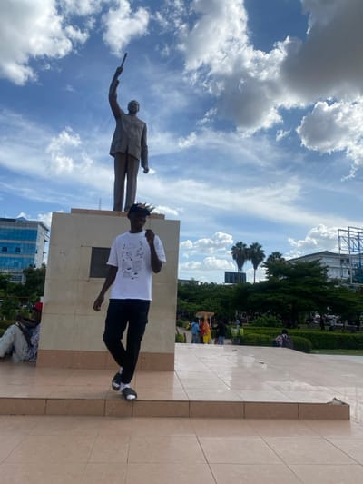 An outdoor image, rotated counter-clockwise, depicts a bright day in what appears to be a public square or memorial park in Ihumwa, Tanzania. The central figure is a young man with dark, spiky hair, wearing a white short-sleeved t-shirt with a black graphic design, dark trousers, and dark sneakers with white soles. He is standing to the right of a prominent bronze-colored statue.
The statue portrays a male figure dressed in a suit, standing upright, and holding an elongated object, possibly a scroll or baton, in his raised right hand. This statue rests on a tall, light-colored stone plinth, which is integrated into the tiled ground where the young man stands. The ground is paved with large, light-colored, possibly beige or tan, square tiles.
In the background, a clear blue sky with scattered fluffy white clouds dominates the upper portion of the frame. Below the sky, lush green trees and manicured bushes suggest a well-maintained park or garden setting. Beyond the greenery, several modern buildings are visible, including one with reflective blue windows, indicating an urban landscape. Several other indistinct figures can be seen scattered among the trees and further back in the park area, some standing, some seated. In the lower right corner, a person wearing a red cap and light-colored clothing is partially visible, crouching or sitting.
The scene conveys a calm, sunny day with bright daylight. The young man appears to be posing, perhaps for a photograph, while others are casually present in the public space. No specific event is clearly identifiable, and there is no clearly legible text visible in the image.