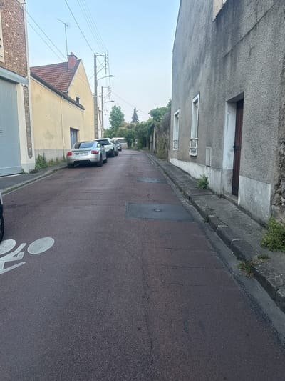 A street scene in Longpont-sur-Orge, France, depicts a narrow road with two parked silver cars and a white SUV. The road surface is a distinctive reddish-brown asphalt, marked with a white bicycle lane symbol. On the left side of the road, a pale yellow building with a red-tiled roof and an antenna is visible, with a light grey garage door. Adjacent to it, another building, rough-textured and grey, features several windows and a dark wooden door. The right side of the road is lined with grey buildings and a concrete curb. The sky is overcast, and trees are visible in the background.