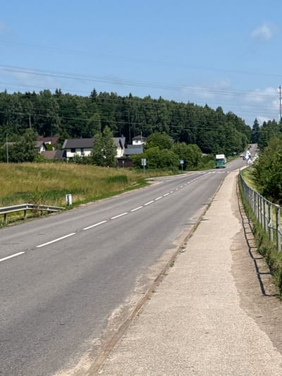 The image depicts a road with a sidewalk in Mažoji Riešė, Lithuania. The road is paved and has white lane markings. A sidewalk runs parallel to the road, separated by a metal fence and vegetation. In the distance, a white and green truck is driving on the road. Several houses and trees are visible on the side of the road, bordering a forest. The sky is clear and blue.
